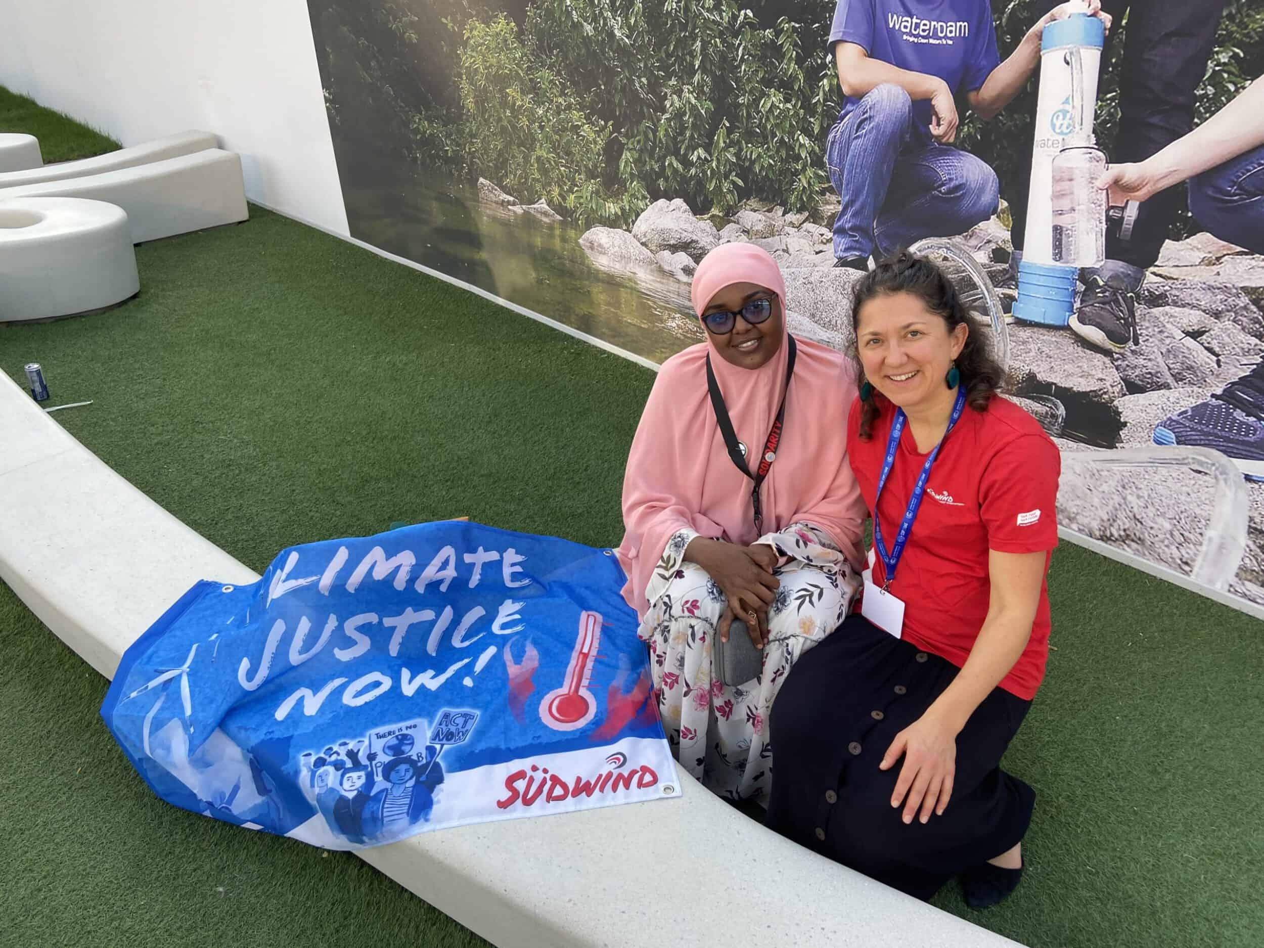 Mana Omar und Lisa Aigelsperger bei der COP28 in Dubai. Neben ihnen ist ein Banner ausgebreitet auf dem steht: "Climate Justice Now"