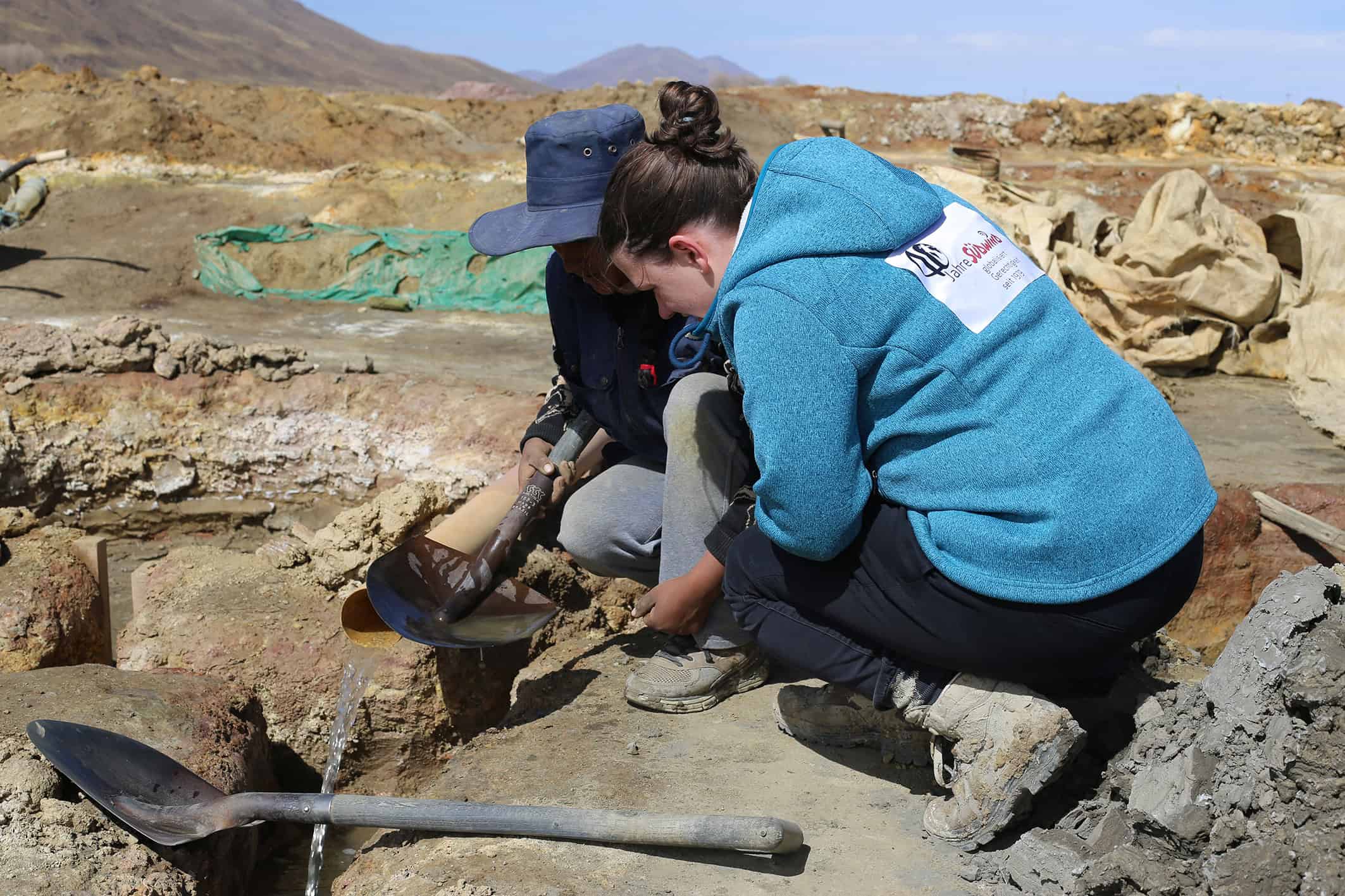 Zinnabbau in Machacamarca in Bolivien: Zwei Personen hocken in einer kargen Gegend auf dem Boden über Werkzeuge gebeugt Zinnabbau in Machacamarca in Bolivien-